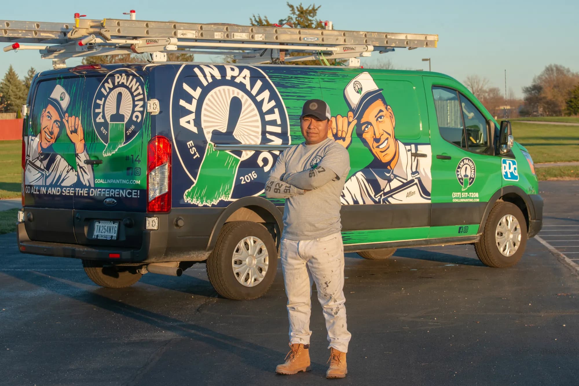 An ALL IN Painting painter in uniform, arms folded, in front of a wrapped ALL IN van with ladders on the roof