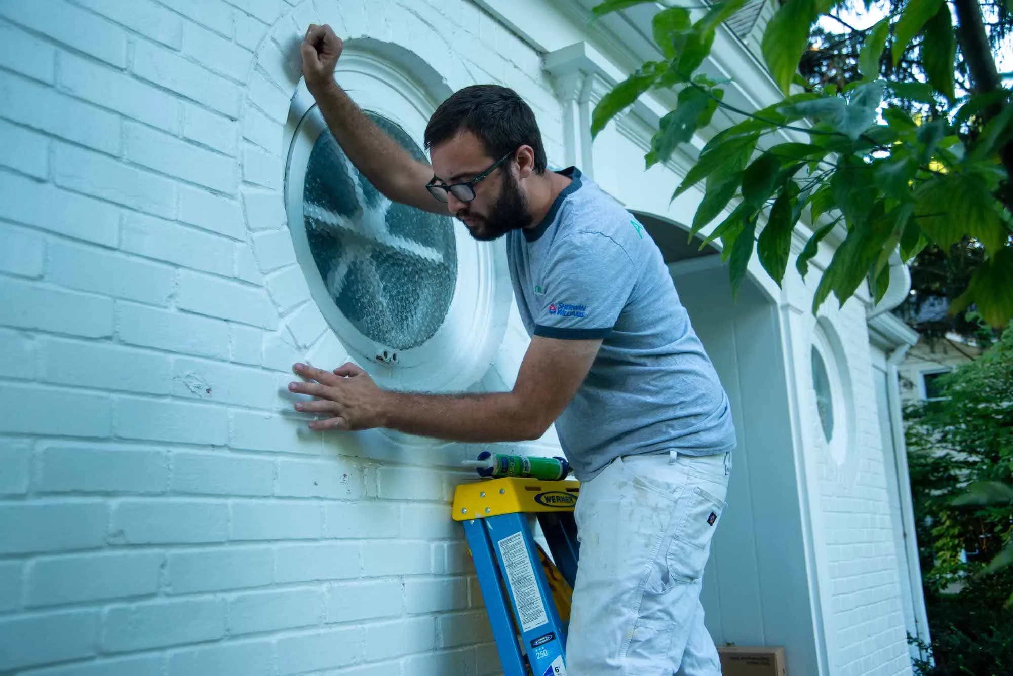An ALL IN painter prepping a round window on white-painted brick